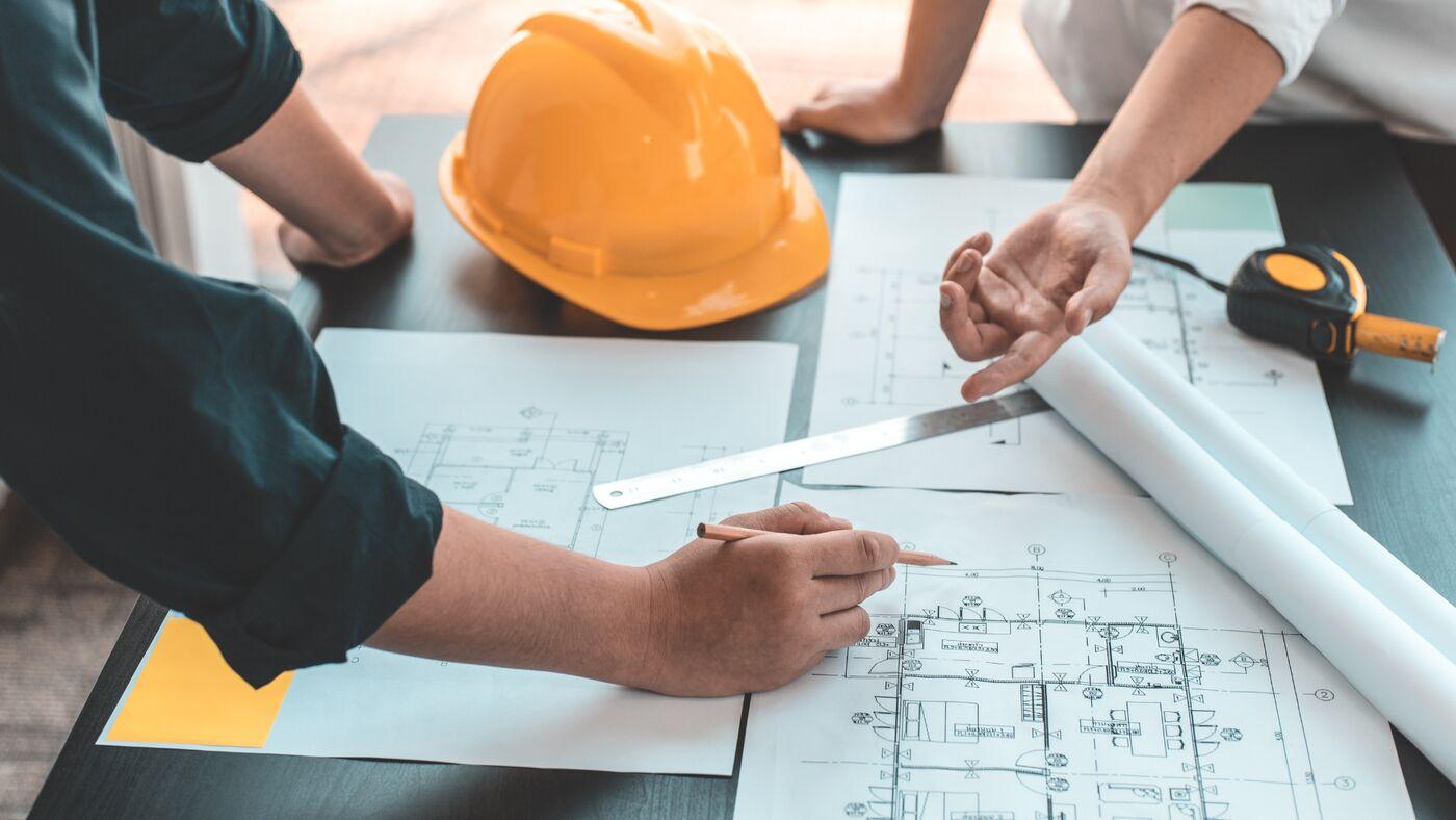 Deux personnes étudient des plans de maison sur une table, à côté d'un casque de chantier jaune, d'une règle et d'un mètre ruban